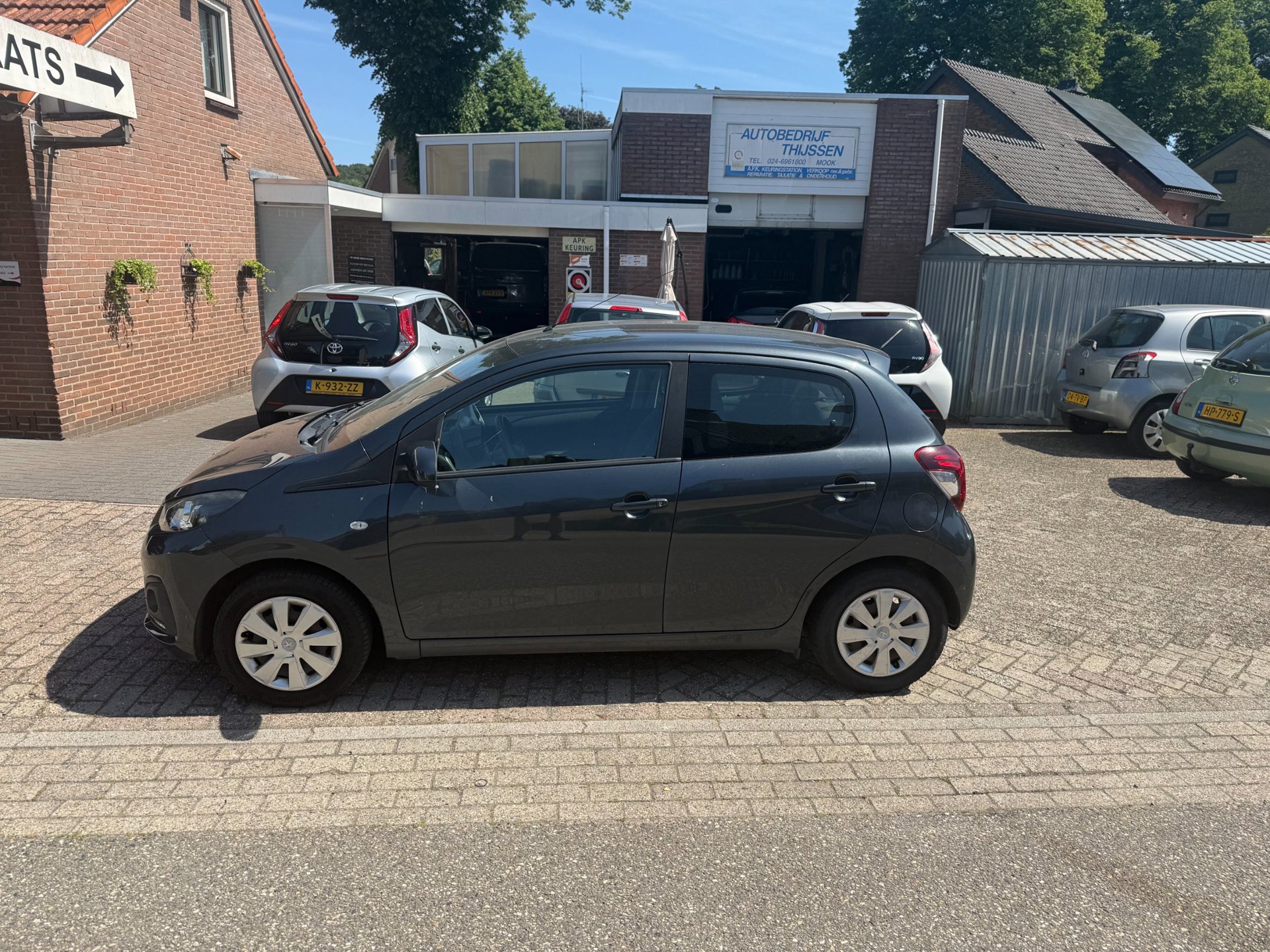 Een donkergrijze Peugeot 108 op het terrein, met de werkplaats erachter