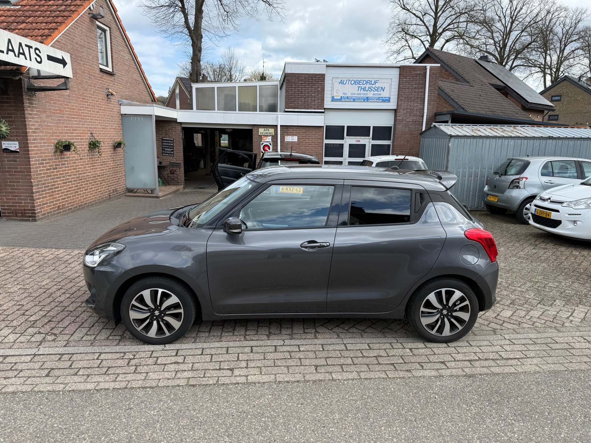 Een grijze Suzuki Swift op het terrein voor de werkplaats van Autobedrijf Thijssen