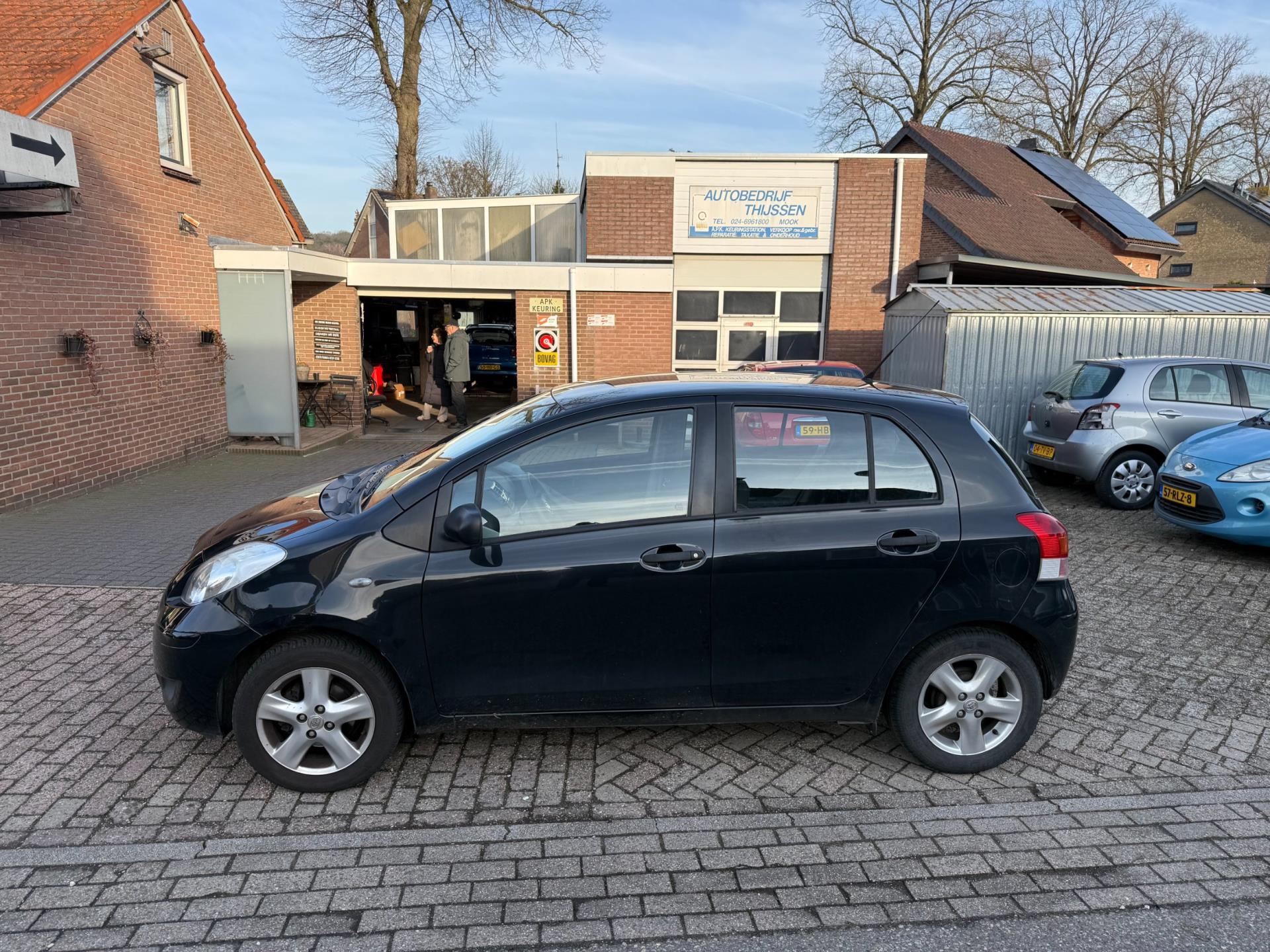 De werkplaats van Autobedrijf Thijssen met geopende werkplaatsdeur en het APK-bord, met een zwarte Toyota Yaris op de voorgrond