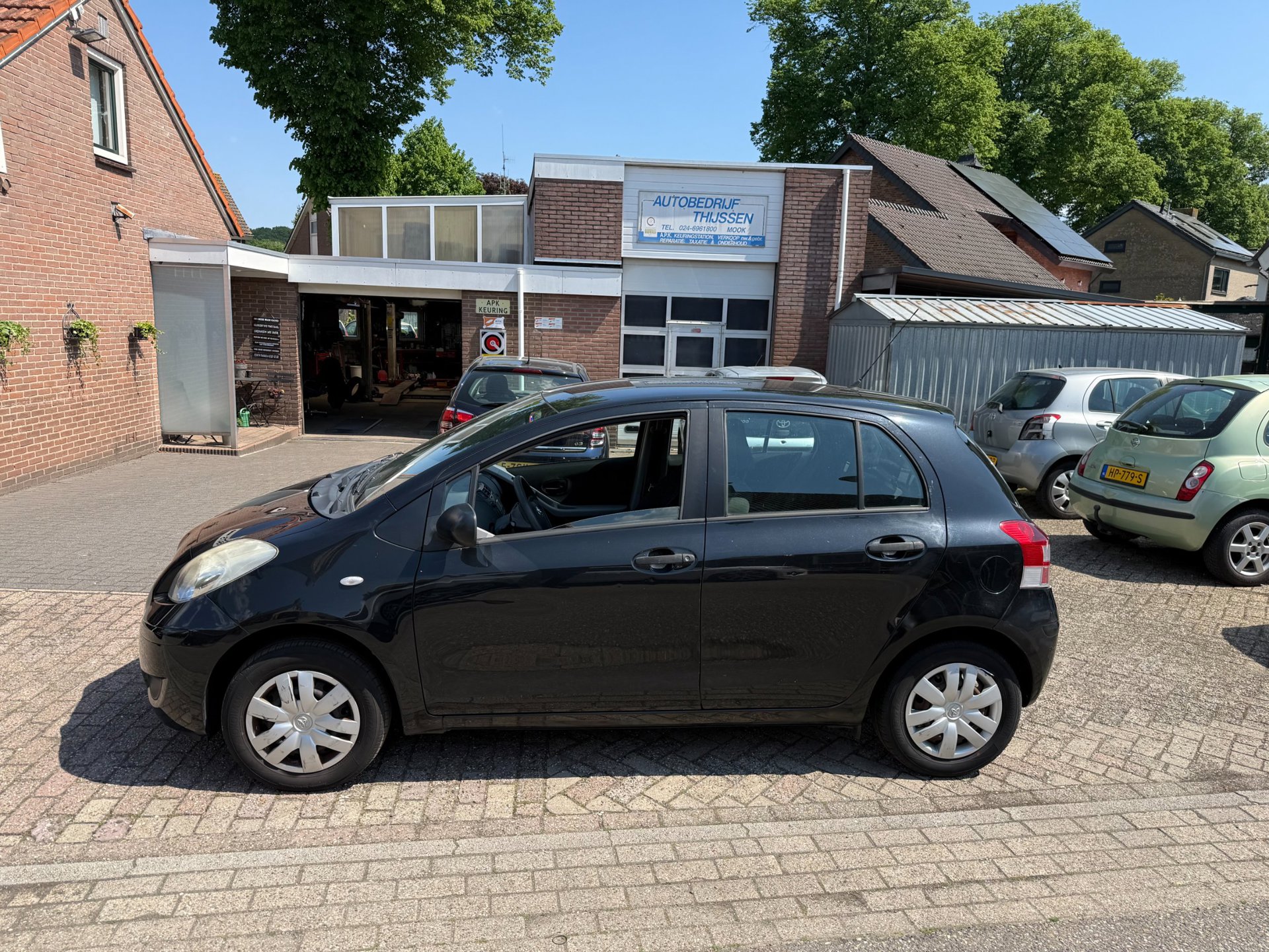 Een zwarte Toyota Yaris voor het pand van Autobedrijf Thijssen, met het bord van het APK-keuringsstation en de werkplaats