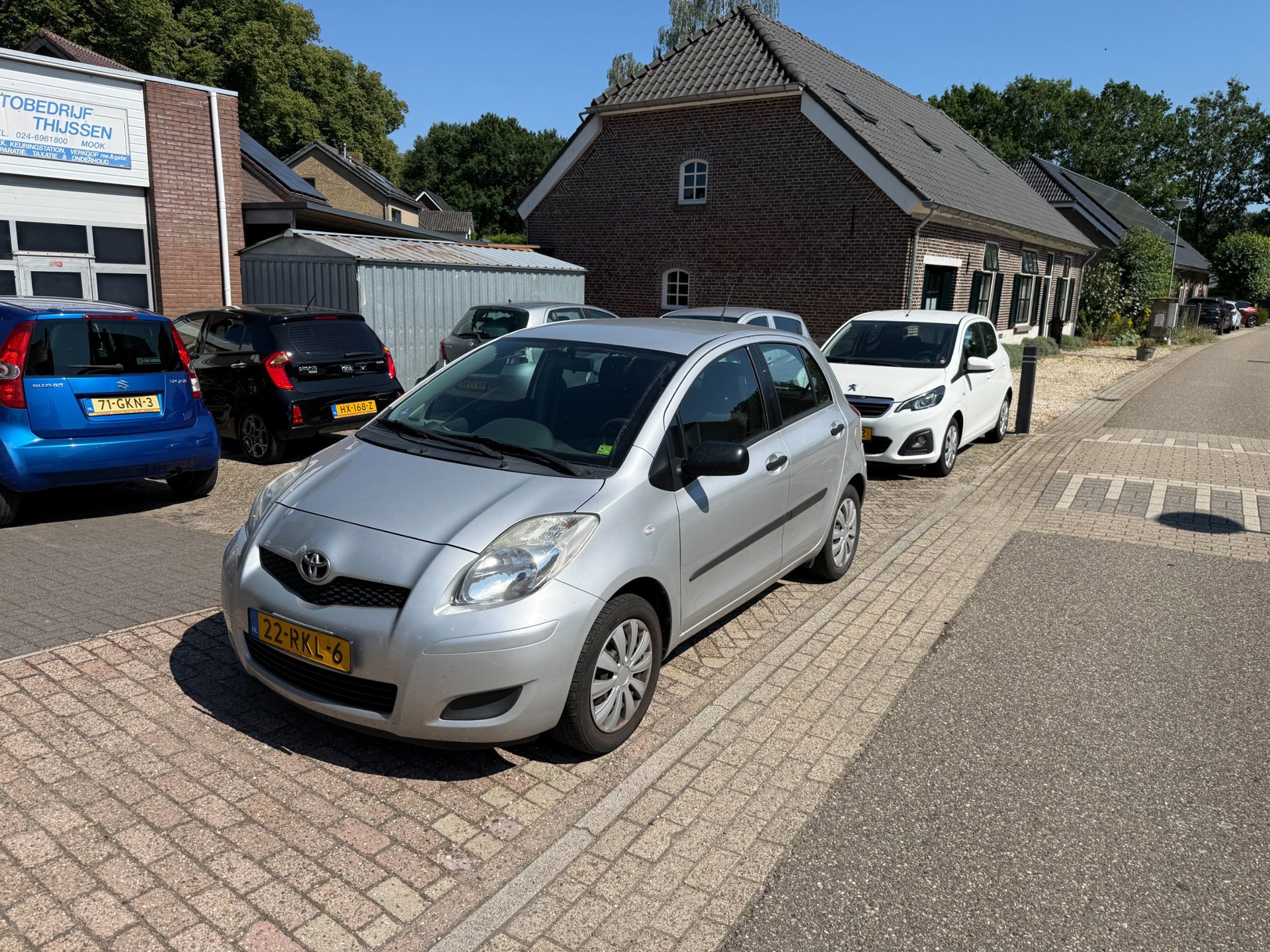 Een zilverkleurige Toyota Yaris op het terrein van Autobedrijf Thijssen, met de werkplaats links achter de auto