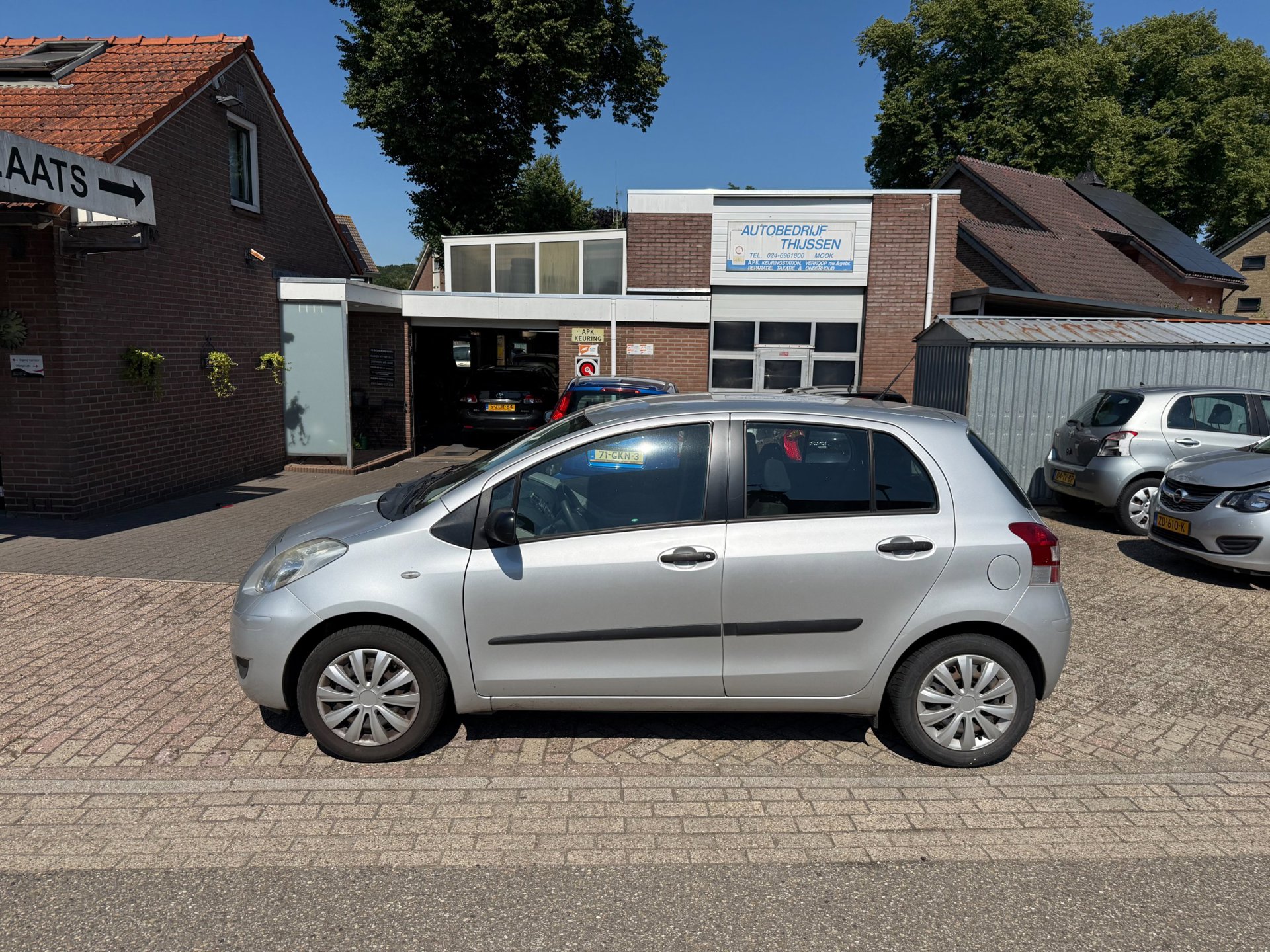 Een zilverkleurige Toyota Yaris op het terrein, met het pand en het APK-bord erachter